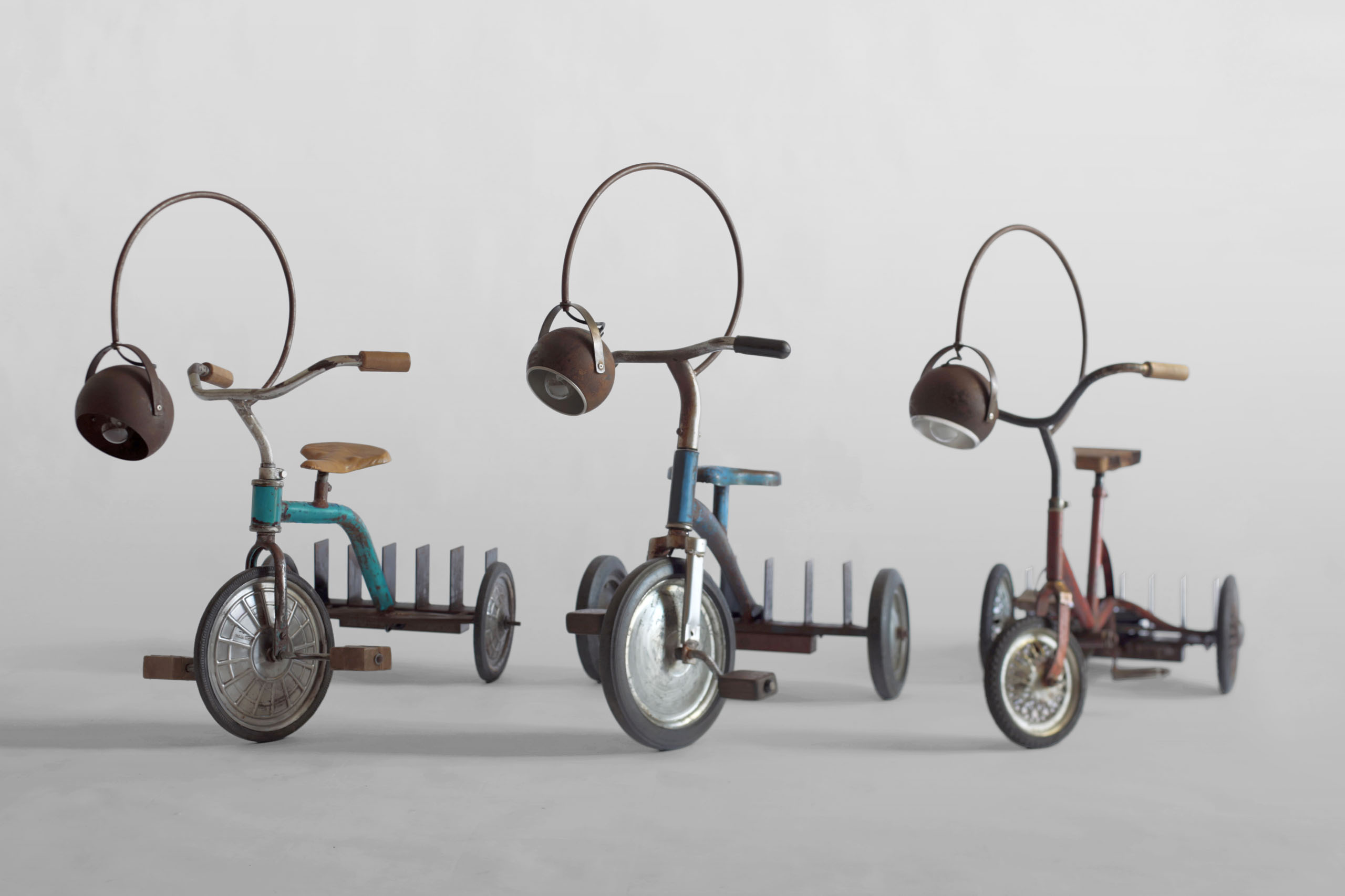 Three original 1960s tricycles — one red, one blue, and one green — transformed into traveling libraries with adjustable reading lamps