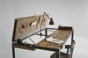 Indoor table shown open as a writing desk with one door raised, red lamp and objects arranged on the wooden top