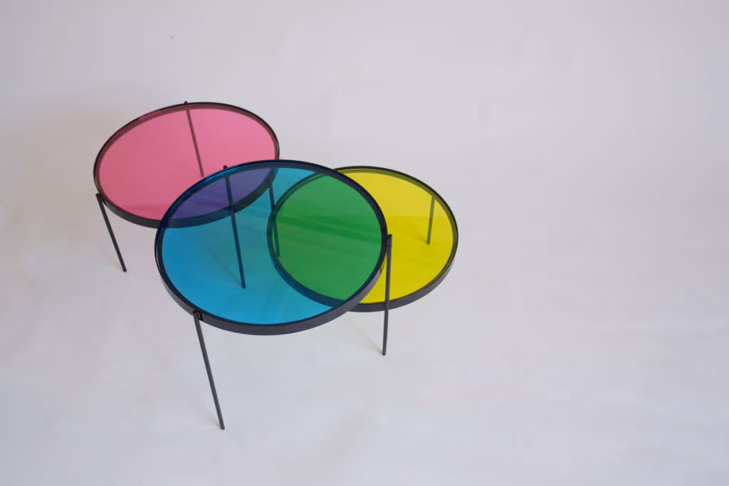 CMYK coffee table by Manoteca, rotation sequence 12: magenta disc continuing rightward rotation, cyan and yellow discs stationary, green intersection fully dominant, magenta nearly disengaged from cyan overlap.