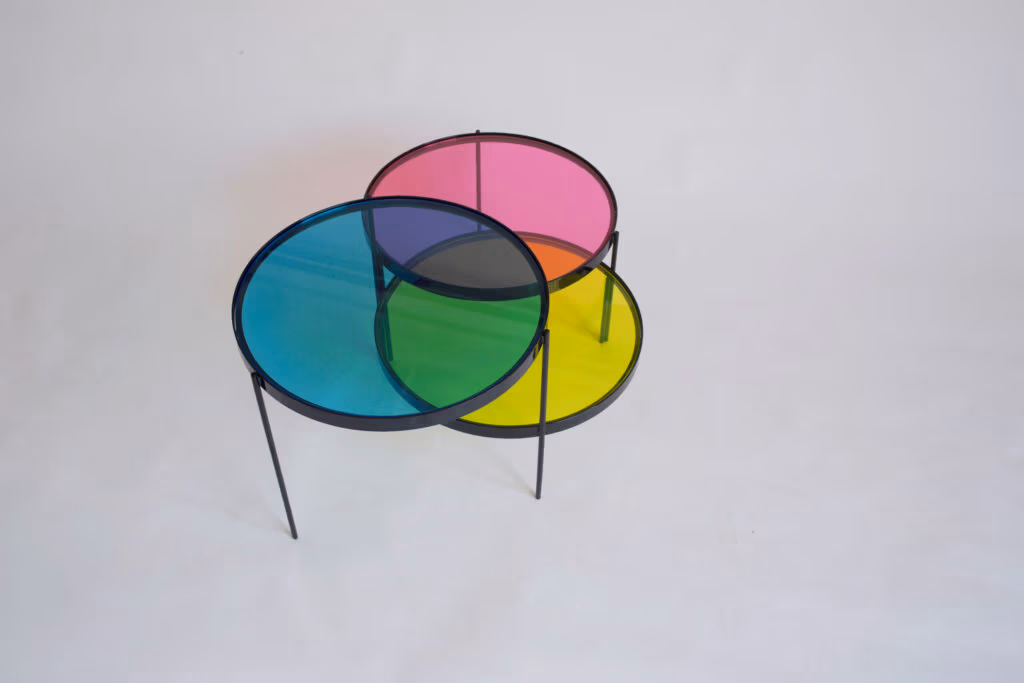 CMYK coffee table by Manoteca, rotation sequence 17: magenta disc continuing leftward, triple overlap at centre reforming, orange magenta-yellow intersection reappearing, cyan and yellow discs stationary.