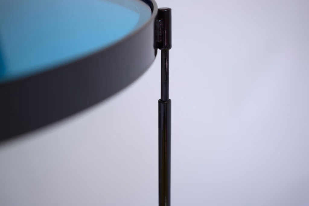 CMYK coffee table by Manoteca, assembly sequence 07: black metal male leg mid-insertion into female socket, cyan glass disc edge in foreground, rotating pin joint forming.