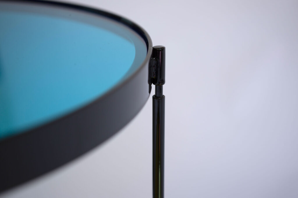 CMYK coffee table by Manoteca, assembly sequence 08: black metal male leg fully inserted into female socket on cyan disc, rotating pin joint complete, cyan glass surface visible.