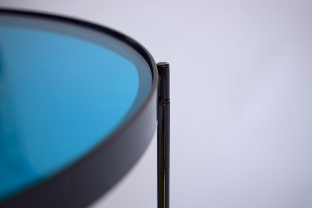 CMYK coffee table by Manoteca, assembly sequence 09: black metal male leg fully inserted into female socket, cyan glass disc surface and black rim in close-up, pin joint detail.